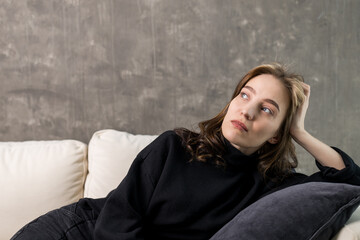 a young woman dreamily looks away lying on the sofa against a gray wall