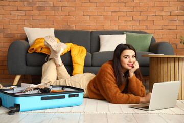 Young woman with laptop packing suitcase at home