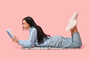Beautiful young woman lying on pink background and reading book