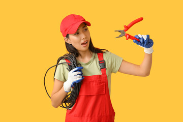 Young female electrician with pliers and cables on yellow background