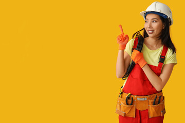 Young female electrician with cables pointing at something on yellow background