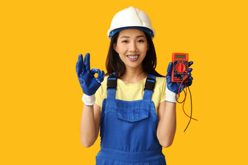 Young female electrician with multimeter showing OK gesture on yellow background