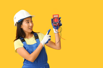 Young female electrician pointing at multimeter on yellow background