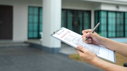 Foreman's hand is using a pen checking on building inspection report form to QC building quality of the house (background). Construction industrial working scene concept. Close-up and selective focus.