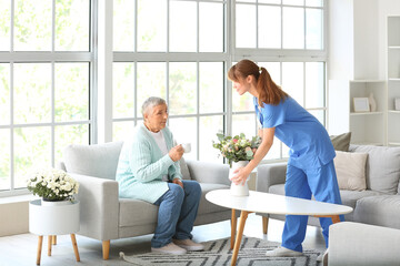 Senior woman and caregiver putting bouquet of flowers on table at home - obrazy, fototapety, plakaty