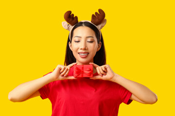 Happy young Asian woman in reindeer horns with Christmas gift box on yellow background