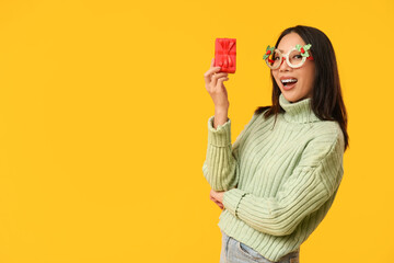 Young Asian woman in decorative eyeglasses with Christmas gift box on yellow background