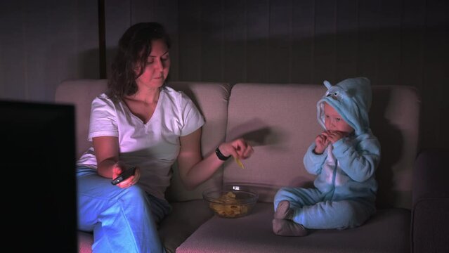 Mom and little daughter in pajamas sit on the sofa at night and watch TV and eat chips. High quality 4k footage