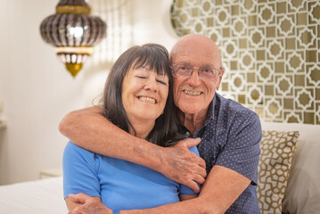 Happy relaxed senior couple sitting on bed hugging and looking at camera. Smiling elderly man and woman enjoying cuddle moments
