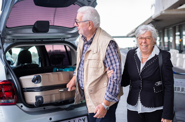 Smiling elderly couple ready for a vacation unload their suitcases from the car outside the...