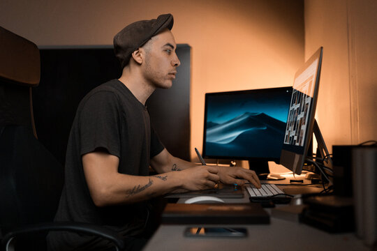 A professional creative retoucher or designer working in a dim studio, using a graphics tablet and stylus to edit photos on a large desktop monitor with a focused expression.