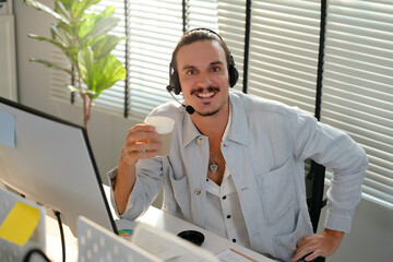 Portrait of Hispanic office worker sitting at desk with headset while holding a cup of hot coffee, smiling face when finish communication. Call center agent support customer to provide information.