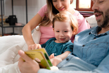 Father reading a bedtime story to his son at home, kid's eye focused on colorful cartoons in book while mother hugs a child.