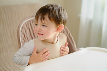 The child is impatiently waiting for food sitting at the table in a bib. Kid boy aged two years (two-year-old)