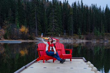 Senior woman traveler with white sweater sitting on red chair at Pyramid Lake, Banff city, Canada. Happiness lady taking selfie with mobile and smiling while traveling alone, tourist, vacation