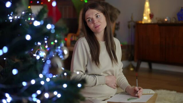 Pregnant woman drawing while sitting near Christmas tree in lotus position. Leisure on the eve of Christmas and New Year