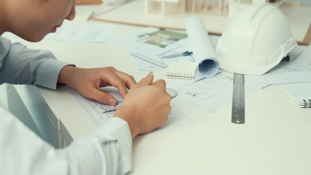 Close up of skilled architect engineer hand draws blueprint on meeting table with blueprint, architectural equipment, house model and safety helmet scatter around. Focus on hand. Immaculate.