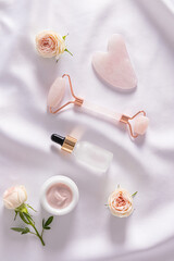 Rose quartz roller massager, gua sha scraper, cosmetic glass matte bottle on delicate white satin. rose flower. Beauty concept. . Top Vertical view.