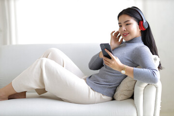 Obraz premium Young asian woman using smartphone and wearing headphone sitting on couch at home. Happy girl relaxing comfortable in the living room, spending lazy weekend, listening to music, watching movie.
