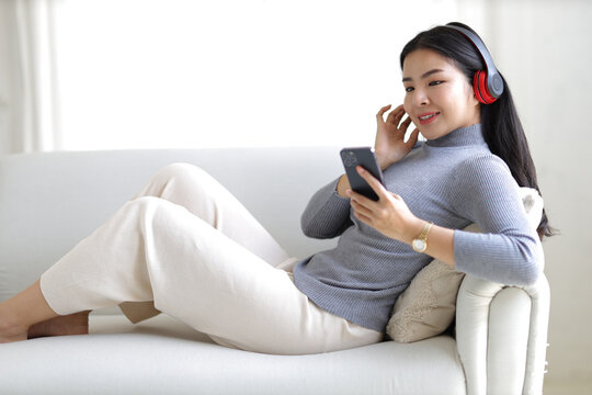 Young asian woman using smartphone and wearing headphone sitting on couch at home. Happy girl relaxing comfortable in the living room, spending lazy weekend, listening to music, watching movie.