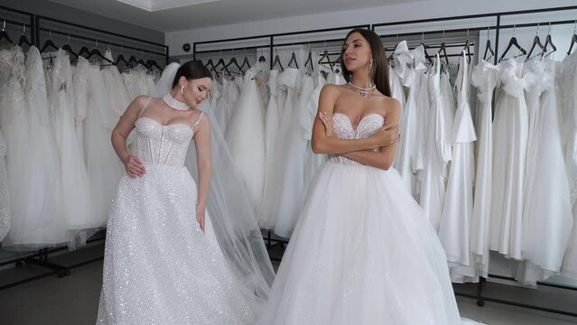 Portrait of two gorgeous young women in wedding dresses with jewelry in a wedding salon, they try on dresses and veils. There are a lot of dresses on hangers in the background. Stable image.