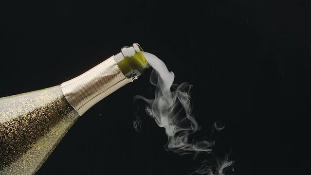 Steam slowly comes out of the neck of an open wine bottle of champagne, followed by champagne foam. Slow motion video, black background.