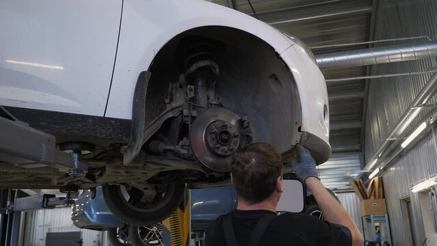 A male mechanic repairs the wheels in a car at a service station, he removes or puts on the fenders in a car that is on a lift. Repair of wheels in the garage.