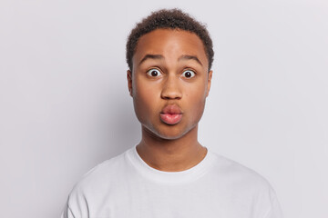 Photo of surprised African man with curly hair keeps lips folded and eyes windely opened cannot believe in shocking news dressed in casual t shirt isolated over white wall reacts to something shocking - obrazy, fototapety, plakaty