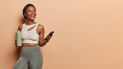 Horizontal shot of athletic dark skinned woman poses with bottle for sport and smartphone browses fitness app looks gladfully aside poses against brown background copy space for your promotion