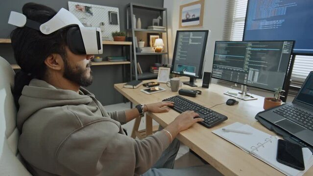 Medium shot of young male software developer in sweatshirt, with beard sitting at desk in home office, writing computer code for dimensional model, and looking at result with VR goggles