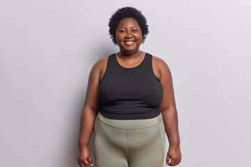 Indoor shot of dark skinned overweight woman smiles happily satisfied after workout at gym dressed in black top and leggings isolated on white background. Curly haired African female goes in for sport - obrazy, fototapety, plakaty