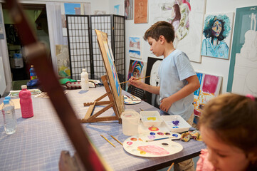 Confident focused boy and girl, artists painters standing by wooden easel and learning art in a...