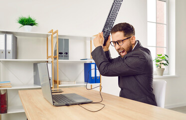 Portrait of an angry shouting business man working on his workplace in office trying to destroy the monitor with his keyboard feeling frustration from work. Overwork, stress concept. - obrazy, fototapety, plakaty