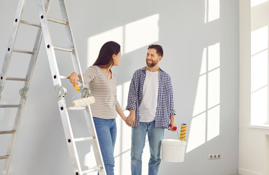 Happy young couple painting house walls in new apartment. Loving spouses looking at each other and holding hands. Smiling husband and wife with paint rollers renovating and decorating room