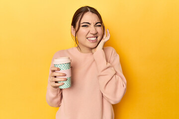 Young caucasian woman holding a takeaway coffee cup covering ears with hands.