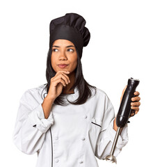 Young Filipina chef with hat holding a mixer looking sideways with doubtful and skeptical...