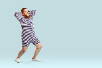 Funny fat man in swimwear having fun on the beach during summer holiday. Happy cheerful excited plump young guy wearing striped swimsuit or underwear posing on light blue copy space studio background - obrazy, fototapety, plakaty