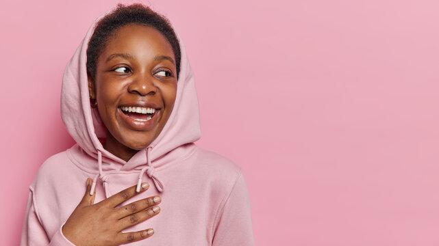 Happy dark skinned millennial girl keeps hand on chest and smiles broadly laughs at something funny concentrated aside wears casual sweatshirt isolated over pink background copy space on right
