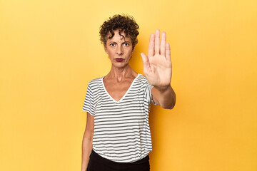 Mid-aged caucasian woman on vibrant yellow standing with outstretched hand showing stop sign, preventing you.