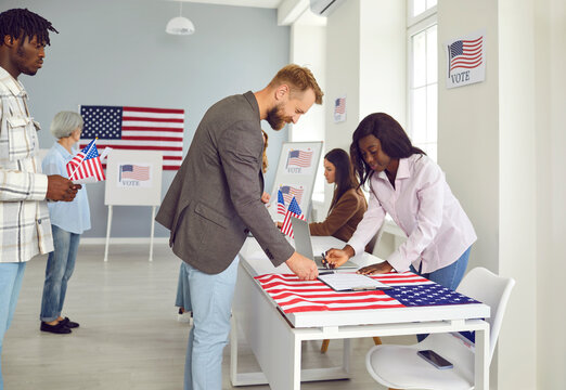 Diverse citizens come to vote on election day in USA. Happy Caucasian male voter standing by registration table with African American female worker at polling place with booths and stars and stripes
