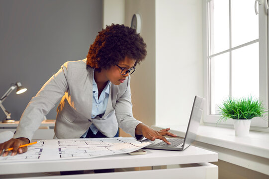 Young interior designer african american woman looking floor plan of new house on laptop monitor screen. Female architect with blueprint construction project standing and working at office.