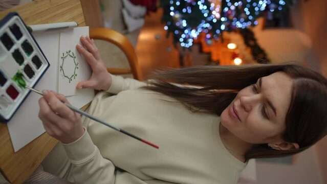 Vertical video. A woman paints a green watercolor Christmas wreath on a card while sitting in a rocking chair against the backdrop of a Christmas tree in a modern home