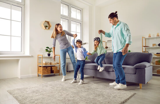 Happy Young Family Playing Together At Home In Living Room. Cheerful Mother And Father Having Fun With Their Kids. Cute Little Boy And Girl Wearing Goggles Jumping On Sofa