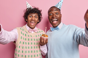Horizontal shot of happy positive men hold cupcake with candle feel positive dressed in festive...