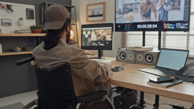 Medium rear shot of young man with disability sitting at desk in wheelchair at home, doing postproduction work on video clips on computer in professional software, while drinking coffee