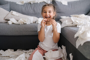 Portrait of little girl eating chocolate at home. Background of scattered napkins. Chaos at home....