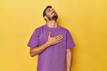 Young Hispanic man on yellow background laughs out loudly keeping hand on chest.