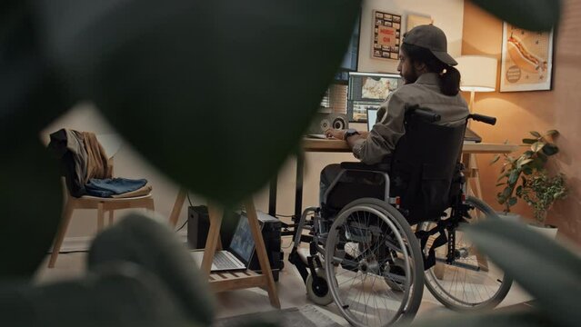 Full rear shot of unrecognizable young man with disability sitting in wheelchair at desk at home, working on computer and editing video clips in professional editing software