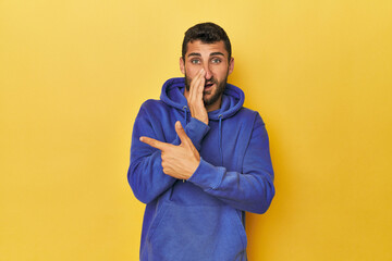 Young Hispanic man on yellow background saying a gossip, pointing to side reporting something.