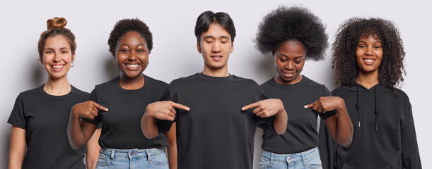 Five diverse people express positive emotions smile gladfully indicates at mock up space of t shirts suggests to write your inscription here stand closely to each other isolated over white background - obrazy, fototapety, plakaty
